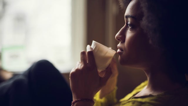 Woman sitting and holding a cup of coffee close to her lips while looking in the distance, thinking ...