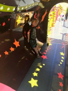 A blonde boy wearing a black shirt and pants standing in a red and yellow tunnel at a playground