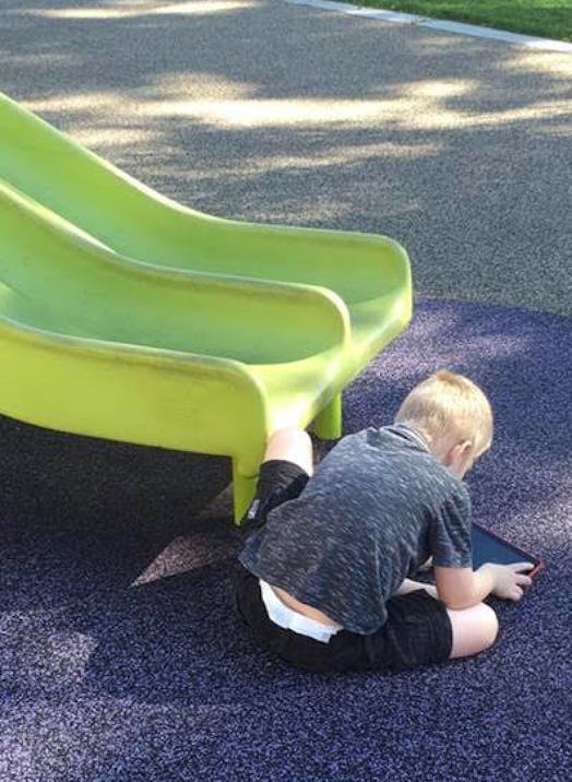 A blonde boy sitting on the ground playing with his tablet underneath a yellow slide on the playgrou...