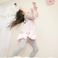 Small girl wearing a pink dress and gray leggings standing on a bed near the curtains and singing wi...