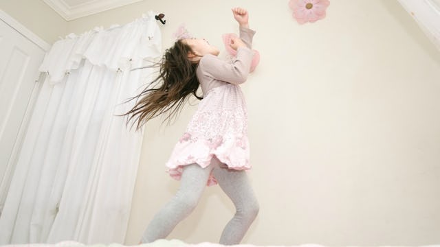 Small girl wearing a pink dress and gray leggings standing on a bed near the curtains and singing wi...