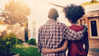 A man in a checked shirt and a woman in a red shirt from the back hugging each other while looking a...