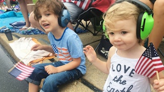 A sensory boy and girl wearing headphones while waving with small American flags and smiling