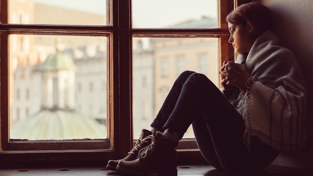 A lonely girl sitting by her window and looking outside, wrapped in a scarf, holding a coffee mug in...