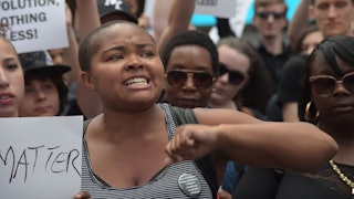 A short-haired woman of color in a grey tank top with a crowd behind her holding a Black Lives Matte...