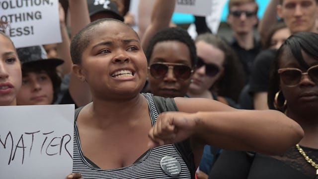 A short-haired woman of color in a grey tank top with a crowd behind her holding a Black Lives Matte...
