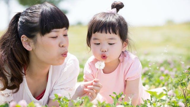 A mother and a daughter in a field sitting and blowing a dandelion puff