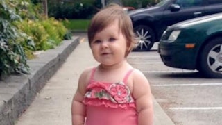 A toddler standing on the sidewalk in a pink dress
