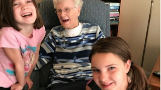 A 95-year-old grandma with glasses in a blue-grey patterned sweater laughing with two of her tween g...