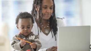 A woman with dreadlocks in a white sweater and a large necklace working on her laptop while her todd...