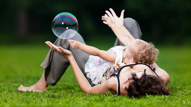 A mother in a black-white tank top and grey pants lying on the grass with her rainbow baby on her re...