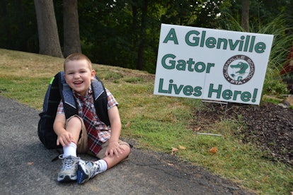 A smiling kid with a backpack sitting on the concrete with "A glenville gator lives here!" sign next...