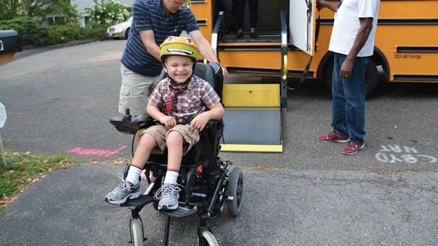 A smiling kid in a wheelchair wearing a square shirt and a Michelangelo hat from TMNT