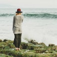 A woman with a hat, standing on a rock, facing the sea and the waves in front of her