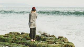 A woman with a hat, standing on a rock, facing the sea and the waves in front of her