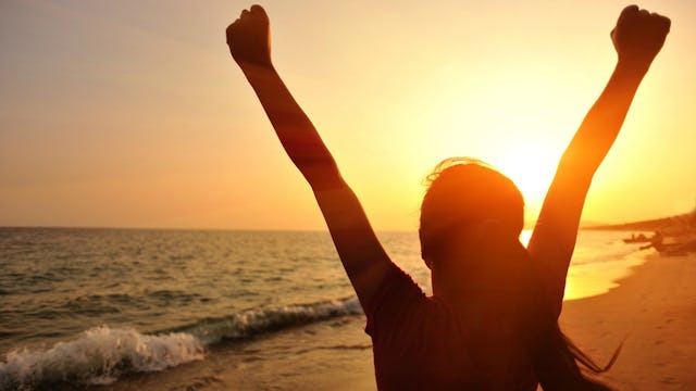 A woman who has overcome childhood trauma standing at the beach with her hands raised in the air.