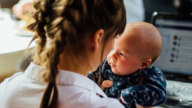 A mom holding a baby and looking at Facebook on her laptop at the same time.