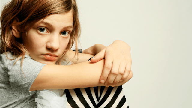 A little girl in a gray shirt and striped leggings sitting down, holding her legs and looking toward...