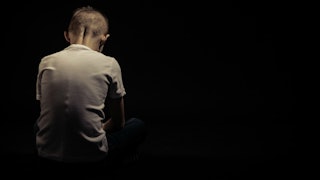 A child in a white shirt sitting in a dark room an grieving after his father's death