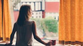 A stay-at-home mom in a white sleeveless shirt waiting for her kids at the window with orange curtai...