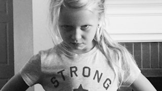 An angry young girl wearing a t-shirt reading 'strong' standing in a room with arms on her hips.