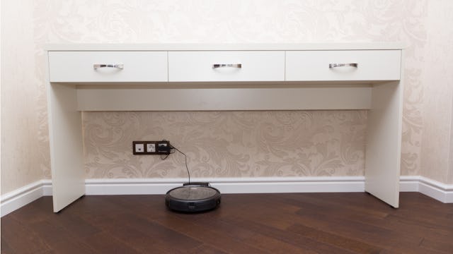 A robot vacuum cleaner on the floor being charged under a white desk