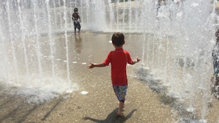 A boy wearing a red shirt standing in a modern fountain between sprinklers and posing with his arms ...
