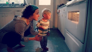 A young mom standing in front of a heated oven with her little son as she's talking about what's bak...