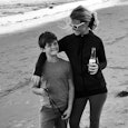Jess Johnston in a turtleneck and sunglasses semi-hugging her child whilst holding a drink on the be...