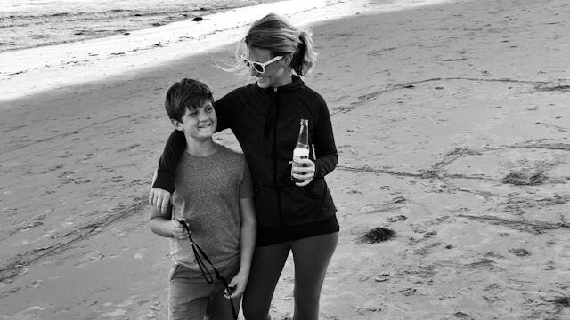 Jess Johnston in a turtleneck and sunglasses semi-hugging her child whilst holding a drink on the be...