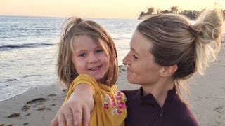 Jess Johnston holding her daughter while looking at her; both are smiling with a beach in the backgr...