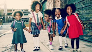 Five schoolgirls on the sidewalk holding hands and smiling at the camera.