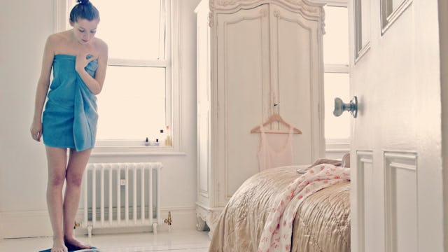 A woman covered with a blue towel looking at a scale in her beige-white color-themed bedroom