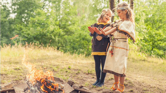 Two Girls In Pocahontas Halloween Costumes Standing Next To A Fire pit In The Forest