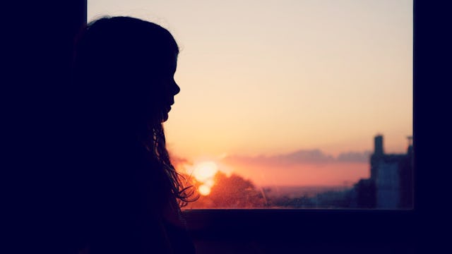 The silhouette of a girl sitting next to a window with a sunset in the background