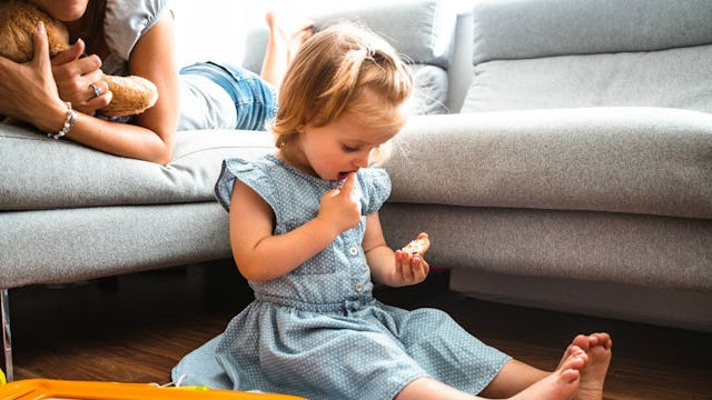 A toddler blonde girl in a light blue dress eating a cookie on the floor while her mother looks at h...