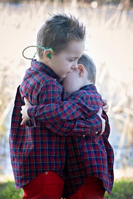 Two young boys wearing the same shirt and red jeans hugging each other while standing on the grass d...