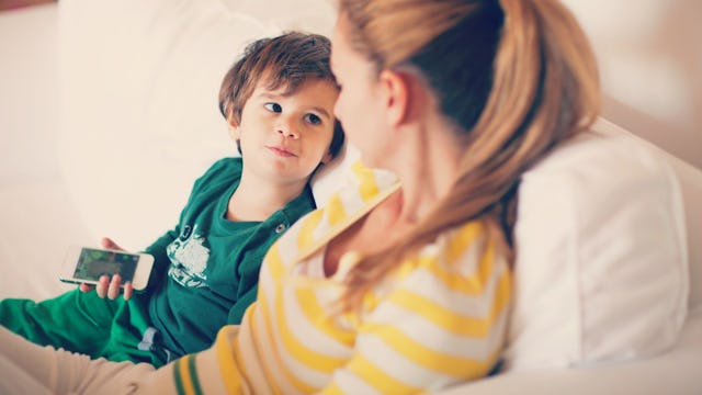 A boy in a green shirt holding a phone about to ask his mom something and they're sitting on a white...
