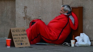 A homeless man in an orange sleeping bag on the street with a sign asking for help next to him