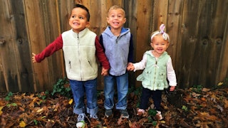 Three smiling toddlers holding hands outdoors.