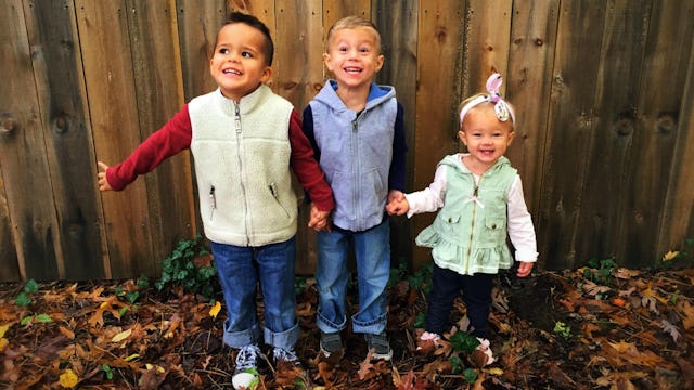 Two toddler boys and a baby girl standing while holding hands, smiling and posing