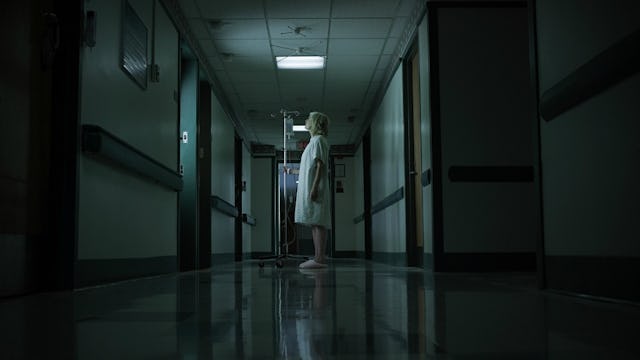 A woman in a dark hospital hall, wearing a hospital gown, suffering from dementia