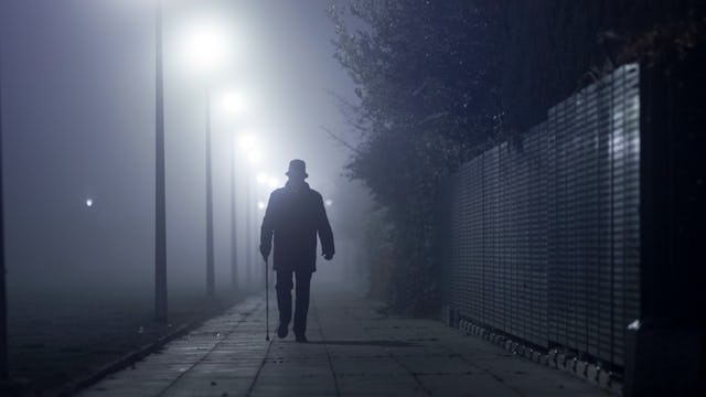 A silhouette of a man with a hat and a cane walking on the sidewalk of an empty, misty street at nig...