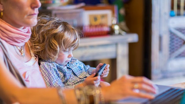 A mother working on her laptop while her child is using her mobile phone and sitting in her lap