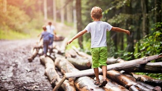 A boy in a white T-shirt and green shorts walking with raised arms on long logs in the forest with f...