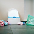 A toddler standing next to their potty and an overturned laundry basket with cloths all over the flo...