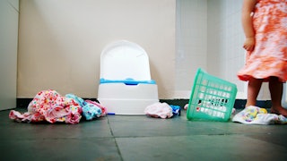 A toddler standing next to their potty and an overturned laundry basket with clothes all over the fl...