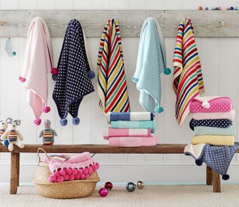 Pink, black, yellow, white, green, and red pom blankets hanging from a wall and folded on a small be...