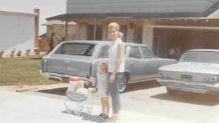 A blonde mother and her daughter holding a kids' toy posing for a picture in front of two old cars p...