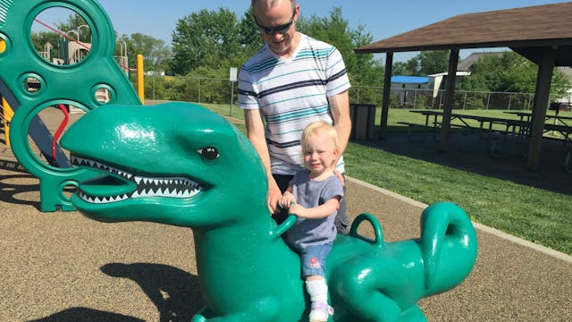 A man in a striped T-shirt playing with his blonde toddler child in a park on a green dinosaur toy i...